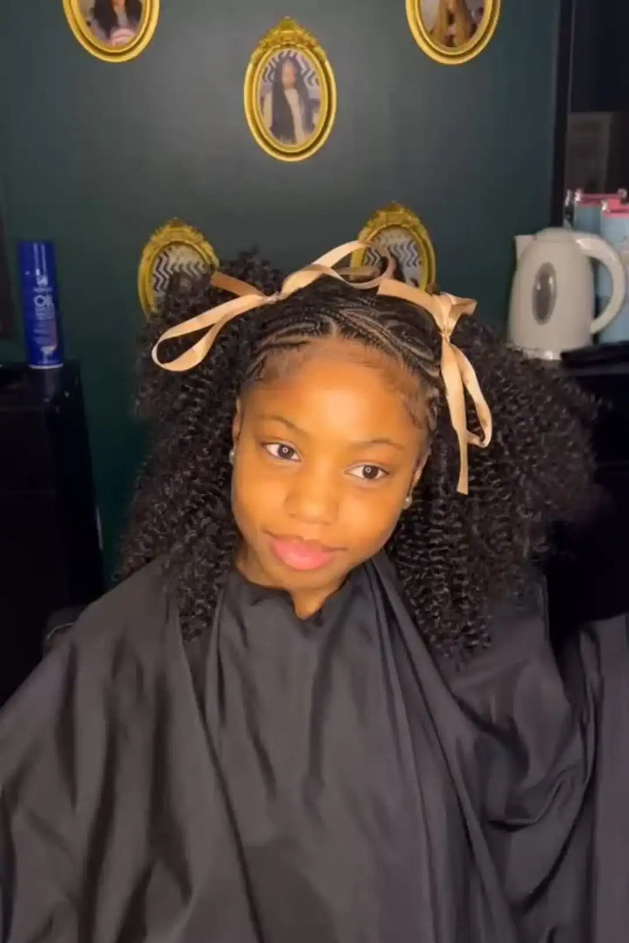 Child with braided hair wearing a black cape in a salon setting.
