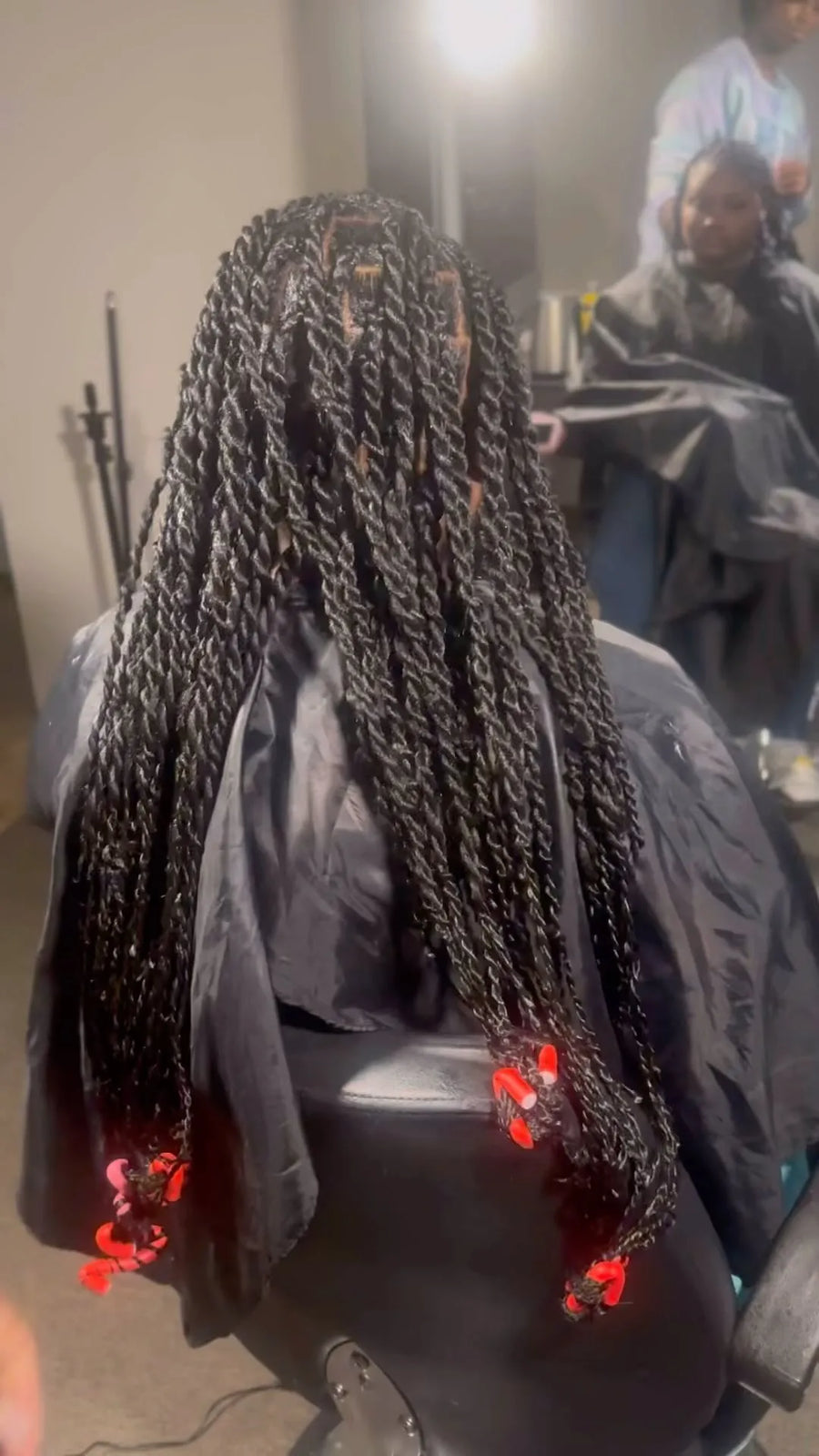 Person with braided hair wearing a black salon cape, with another person in the background.