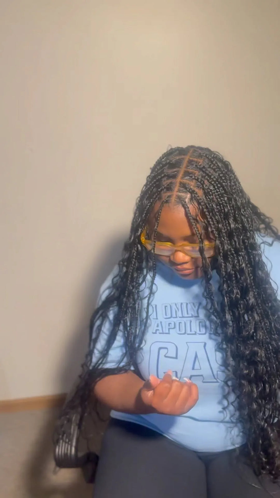 Person with braided hair wearing a blue shirt sitting against a plain wall.