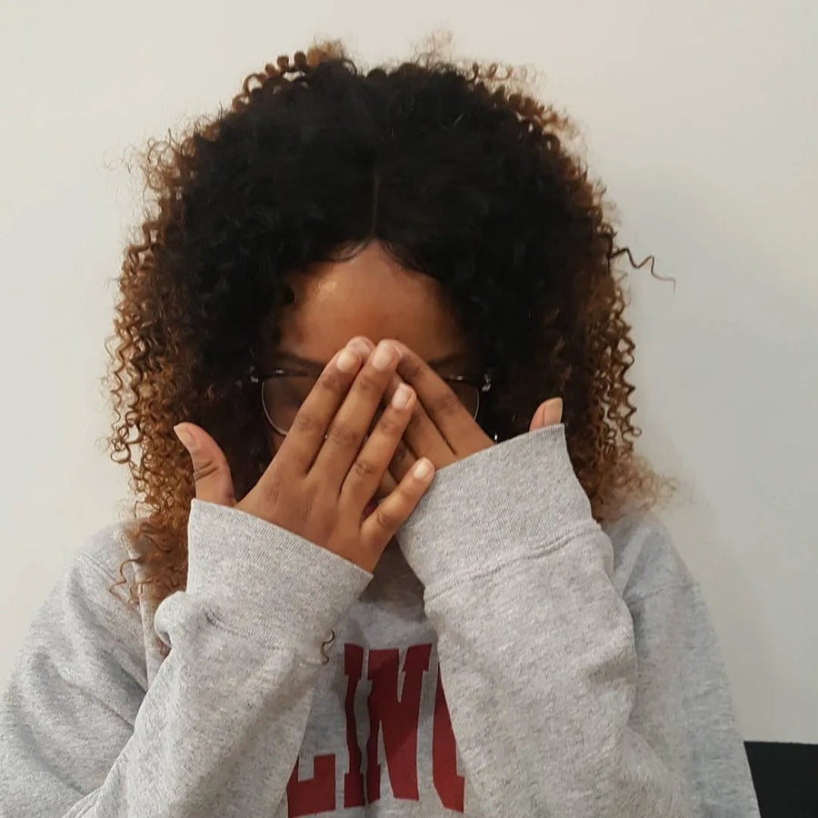 Person wearing a gray sweatshirt with text, covering their face with their hands against a plain background