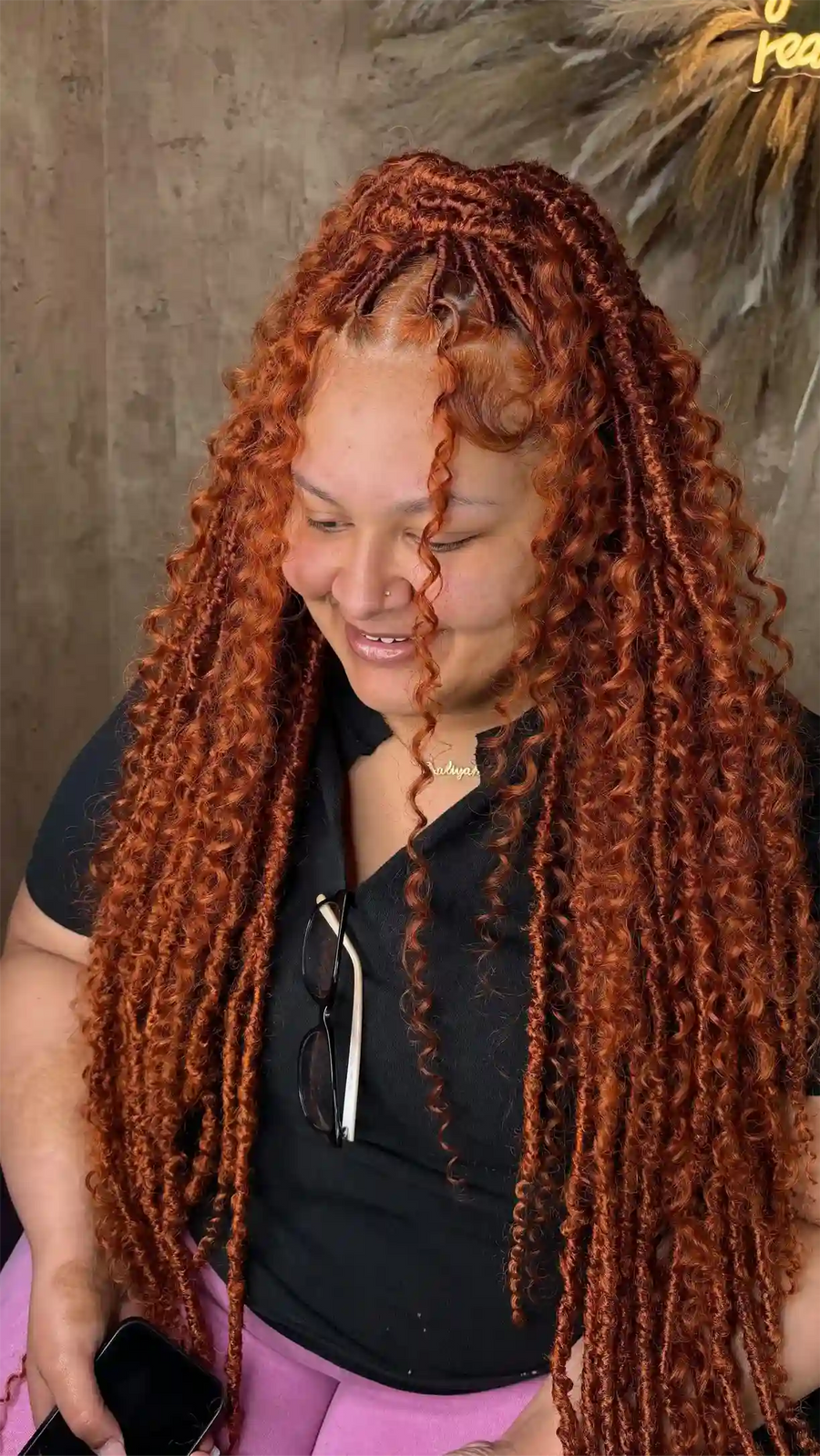 Woman with long, braided red hair wearing a black top and pink pants.