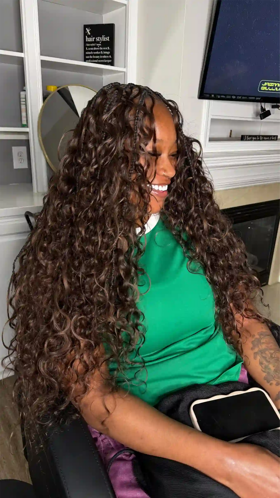 Woman with long, curly hair sitting in a room with shelves and a television.