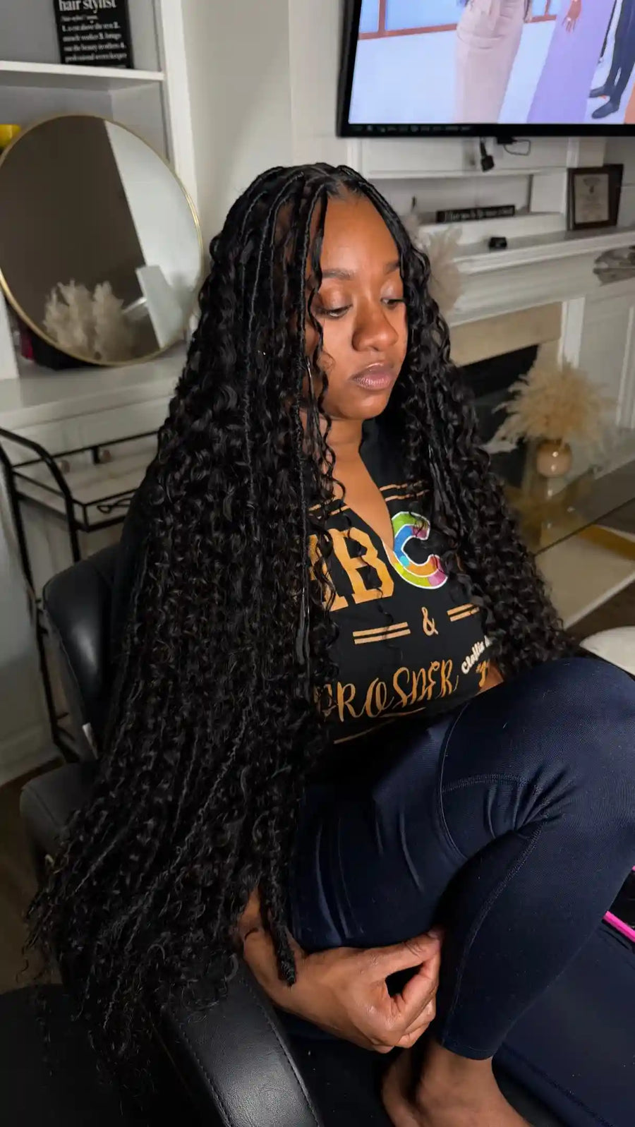 Person with braided hair wearing a black t-shirt with colorful text in a home setting.