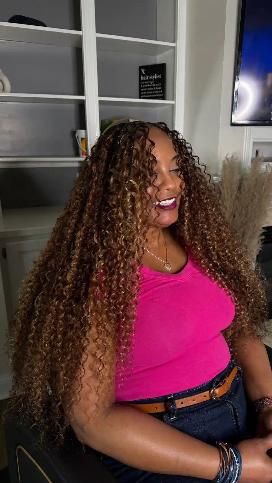 Woman with long, curly hair wearing a pink top sitting in a room.