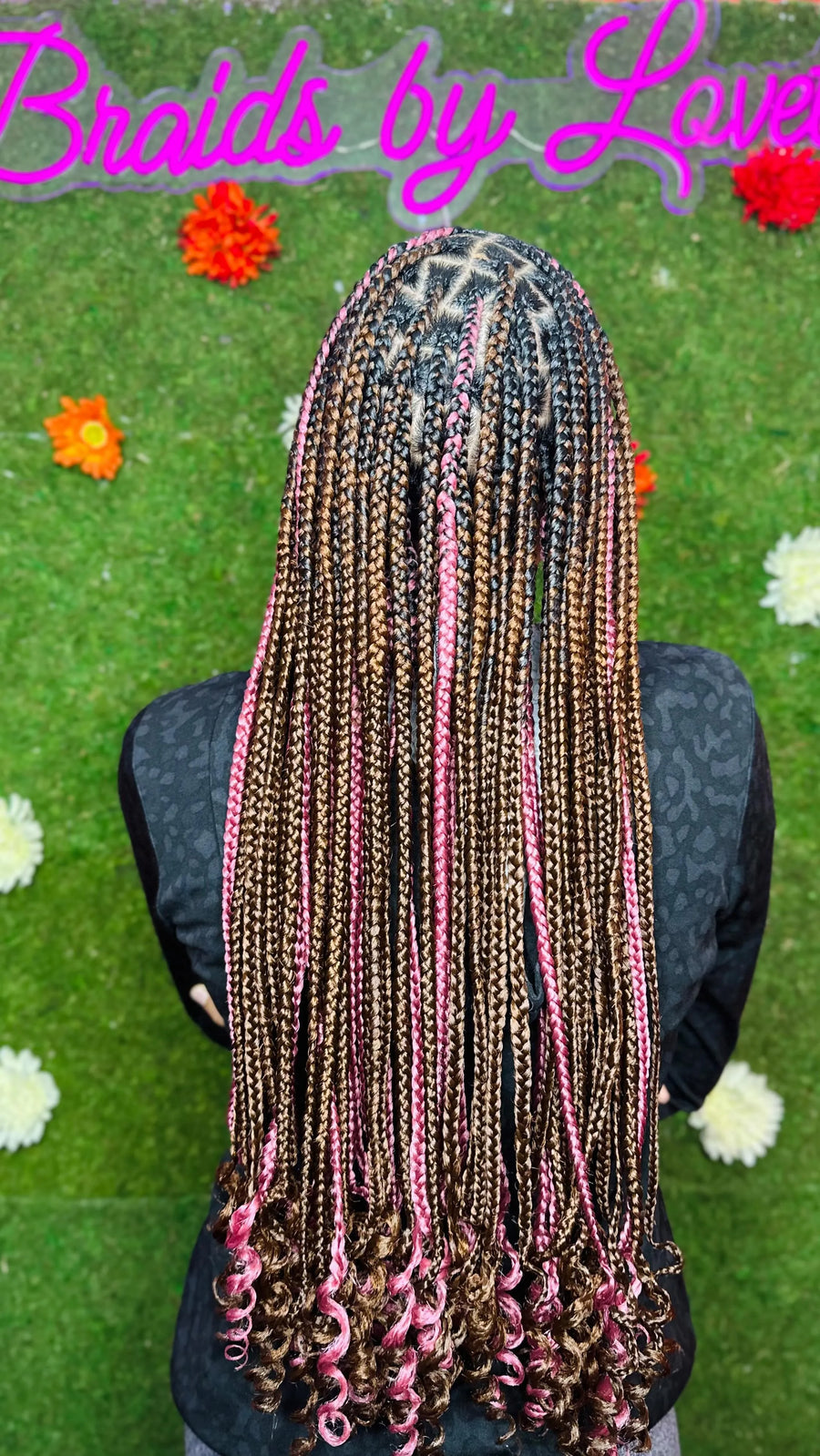Person with braided hair sitting on grass with flowers, featuring 'Braids by Love' text.